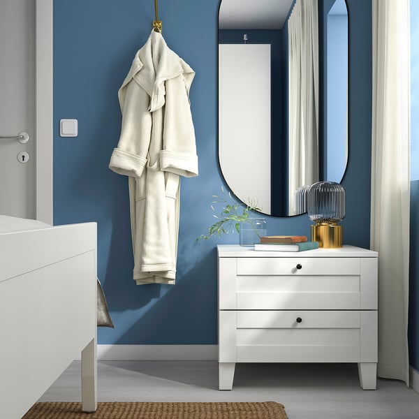Modern bedroom with white SANNIDAL bedside table, blue wall, round mirror, hanging robe, jute rug.