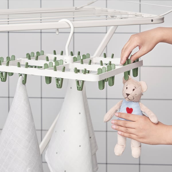 A hand hangs clothes using a white plastic drying rack with 24 green clips. A bear stuffed toy is held nearby.