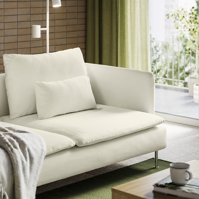A modern white sofa with cushions near a large window covered by green curtains. A cup and book sit on a coffee table nearby.