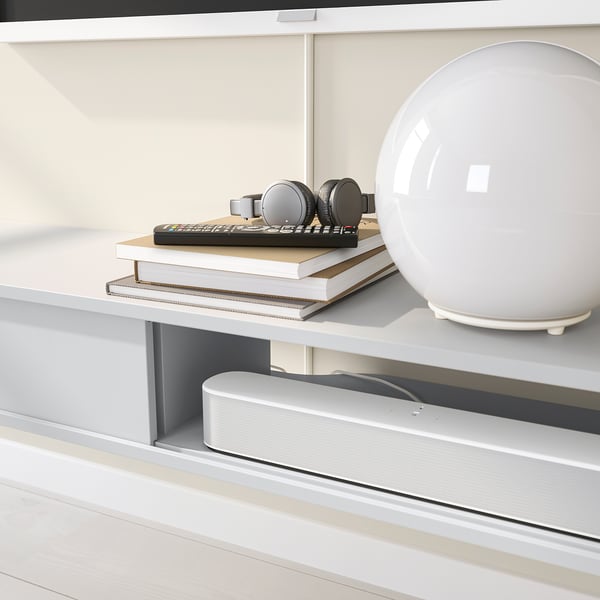 White wall-mounted media shelf with sliding doors hiding cables, holding books, headphones, and a lamp.