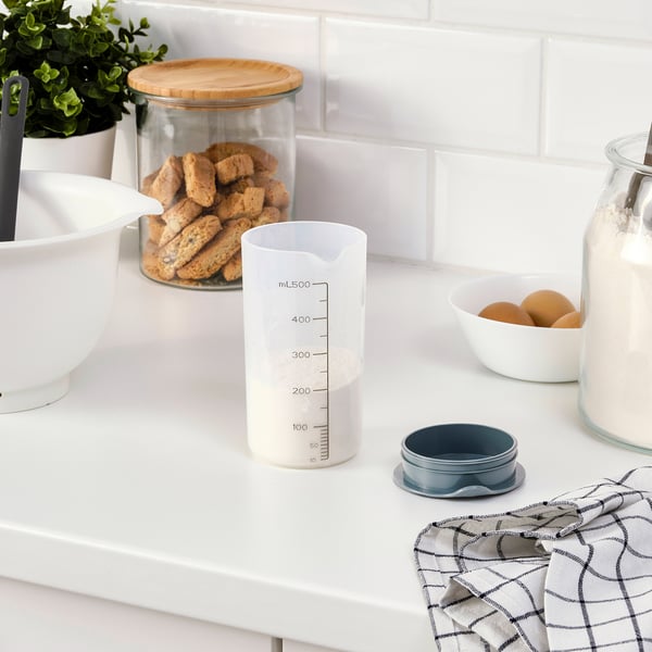 A kitchen countertop has a measuring shaker with a leak-proof lid, containing liquid, with bakeware items around.