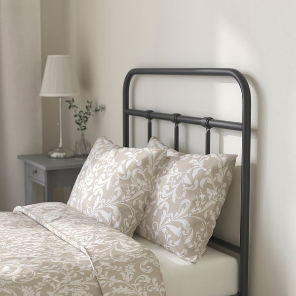 Bed with beige floral bedding, two pillows, grey metal headboard, lamp on bedside table.