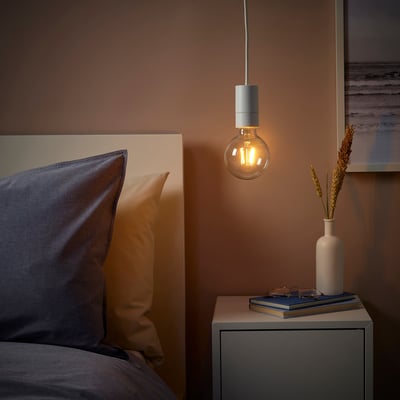 Cosy bedroom with SUNNEBY lamp over bedside table, emitting warm light. Bedside table holds dried plants, books, and glasses.