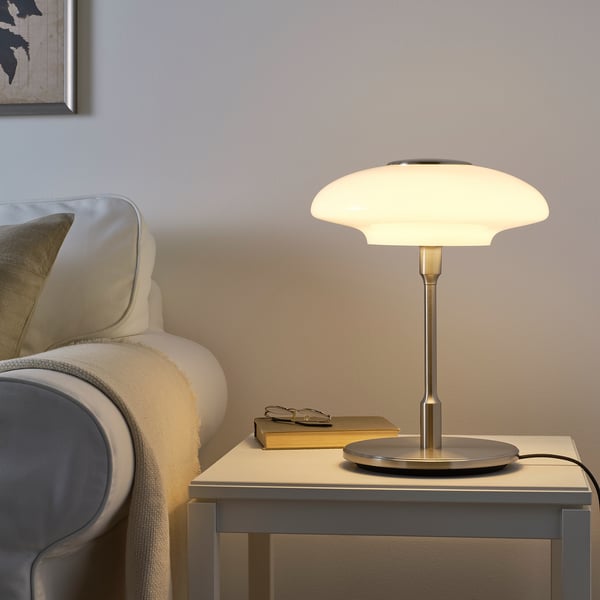 TÄLLBYN lamp on table. Small, white glass shade on metal base. Next to grey and white couch.