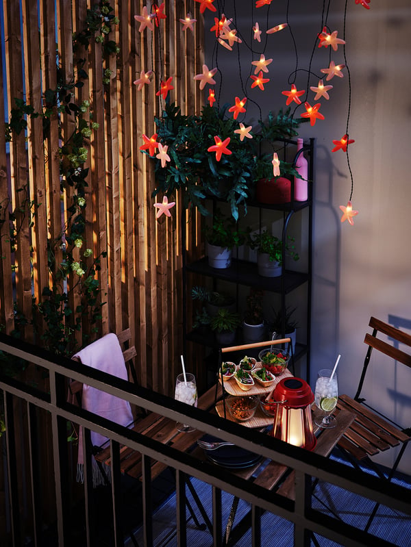 Balcony with TÄRNÖ table and chair, acacia wood, powder-coated steel, lit with star-shaped lights, potted plants, food, drinks.