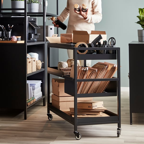 Black TROTTEN trolley with 3 shelves, near shelving units, holding boxes and office supplies.