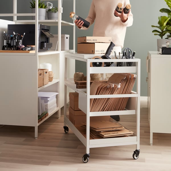 A white TROTTEN trolley with three shelves, holding boxes and office supplies, next to desks.