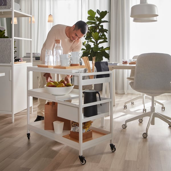 Person organising TROTTEN trolley with fruits, glasses, bottle, and coffeemaker for office storage.