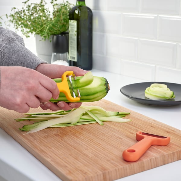 Person peels vegetable with yellow UPPFYLLD cutter on wooden board, orange cutter nearby, producing strips.