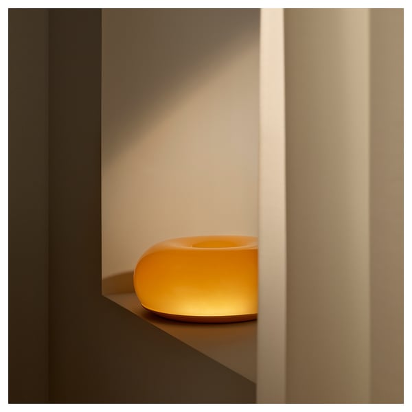 A small, vibrant orange, glass lamp sits on a modern shelf, emitting a warm glow. The donut-shaped design radiates cosy light.