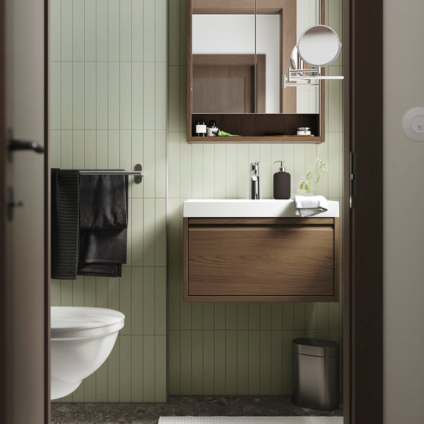 Modern bathroom with light green tiles, white sink, dark wood cabinet, black towels, and metal accents. Vanity fits neatly beneath a large mirror.