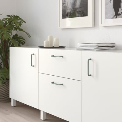White BAGGANÄS sideboard, minimalist design, 2 drawers, 2 doors with silver handles. Between pictures and plant.