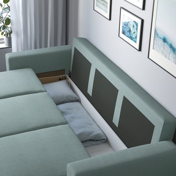 Green sofa-bed with storage open, showing cushions and box inside. Modern living room with art and plants.