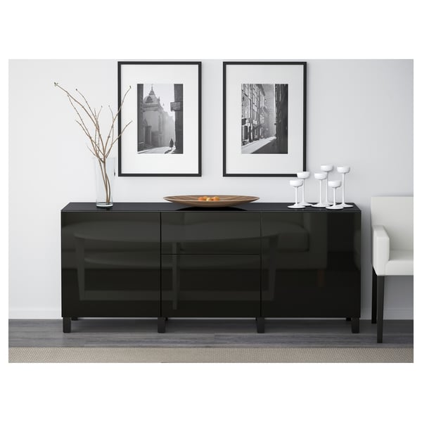 Modern black glass sideboard with drawers and shelves. Topped with decorative bowl and vases, against a white wall with framed photos.