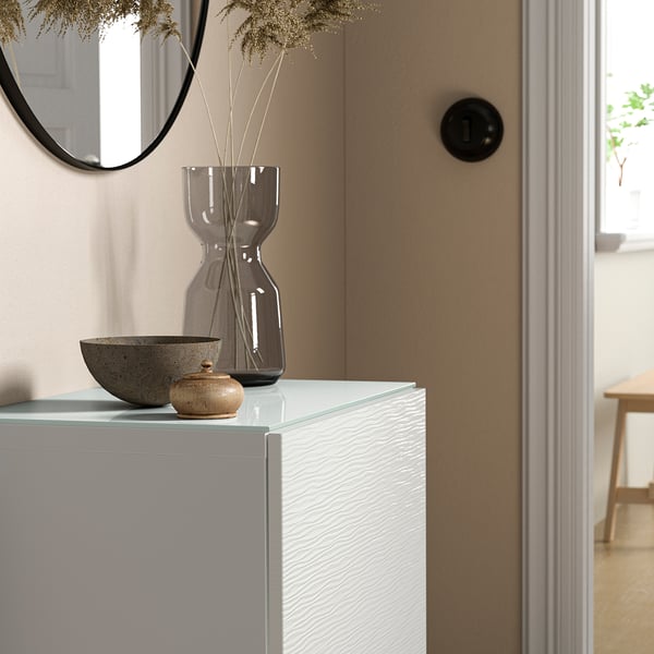 BESTÅ white glass top sits between a vase and a bowl, reflected in a round mirror. Sleek and modern, it complements adjacent wood door and far table.