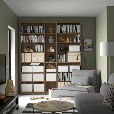 Large wooden BILLY bookcase in corner, holding books & decorations, grey fabric couch beside it.