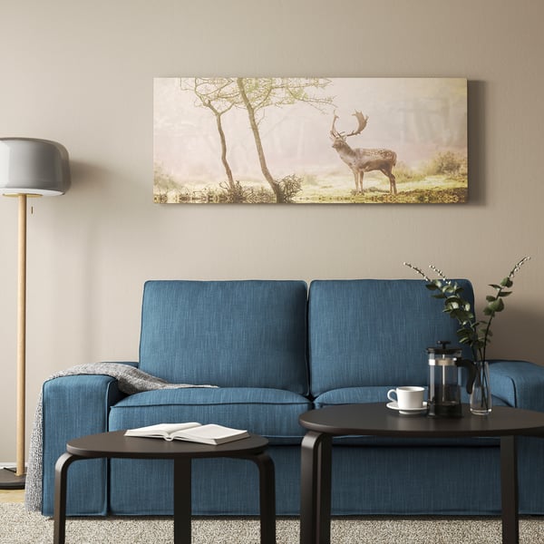 Cosy living room with blue BJÖRKSTA couch, deer canvas, grey lamp, & side table with plants & coffee press.