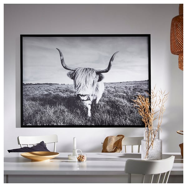 Large framed black and white a bull with long horns in a field on a wall above a white dining table with chairs, vase, and decorations.