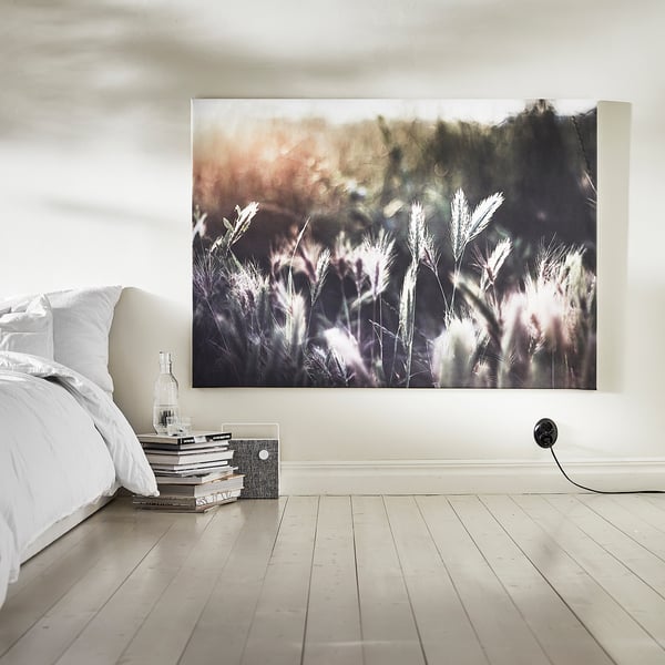 A serene bedroom displays a large BJÖRKSTA picture frame with a nature scene against white walls. A bed with white linens sits to the side.