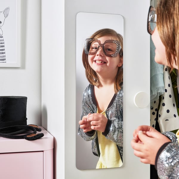 A young girl styles themself in a mirror attached to a wardrobe door.