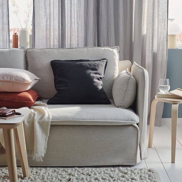 A cosy grey couch with black, grey, and peach pillows, a cream throw, wooden side tables, and a glass of water. Light grey curtains in the background.