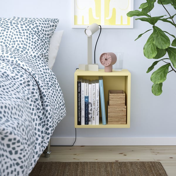 Bedside: yellow EKET cube holds books, clock, glass. Blue bedspread with white pillows. Plant to side. Yellow art above.