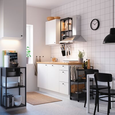 Modern kitchen with white cabinets, black chairs, stainless sink, garlic on shelf, plates on open storage, shelf with plant, and wall clock.