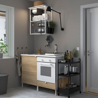 Modern compact kitchen: white oven, wood-panel base, open shelves, black cart with bottles and plants.