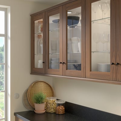 Brown ENKÖPING walnut cabinet with glass doors, housing dishes. Counter holds woven tray, plant, and jars.