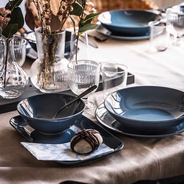 Table set with FÄRGKLAR blue stoneware plates, cups, and clear vases holding flowers and greenery.