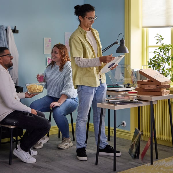 Three people in a cosy blue room. One holds a vinyl, another chips. Two wooden tables with records and food. A lamp lights the scene.