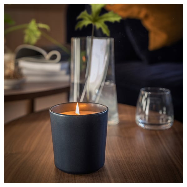 Black ceramic candle, lit, on wooden table with glass tumbler & clear vase.