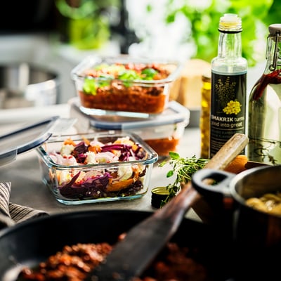 Glass food containers with lids holding colourful salads on a rustic table with a pan and herbs.