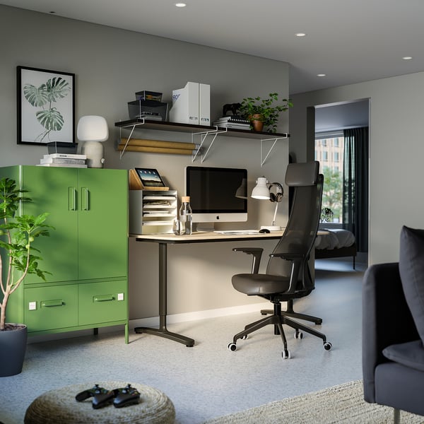 Modern home office with green cabinet, sleek desk, ergonomic black chair, computer, shelves with books, plants, and a grey sofa.