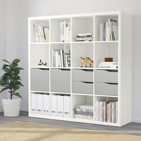 White KALLAX shelf unit with books, binders, decor, and a plant. Customizable storage with shelves and drawers.