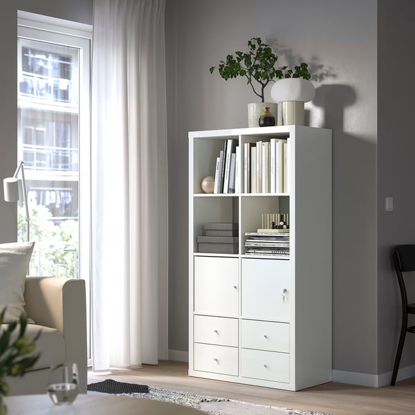 A white KALLAX shelf unit in a bright room with an open shelf displaying books and decorative items, and a cabinet with a lock for secure storage.