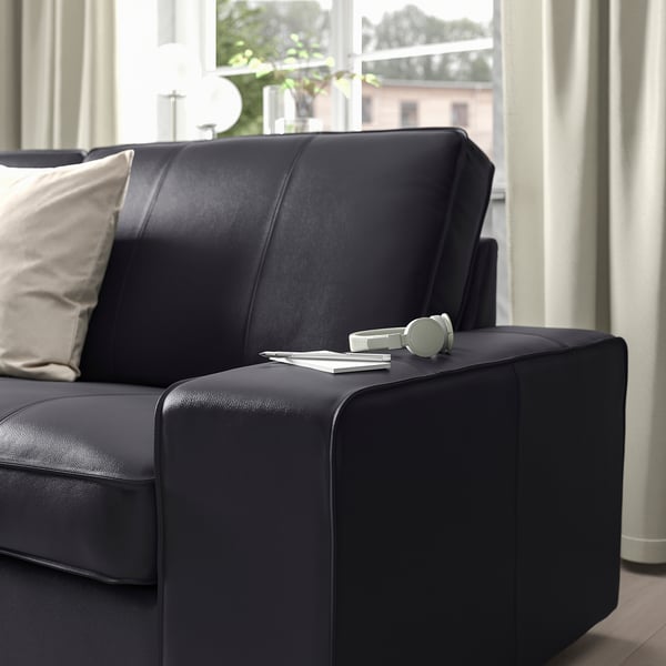 Black leather sofa with armrest, featuring headphones and remote on the armrest, set against a window with curtains.