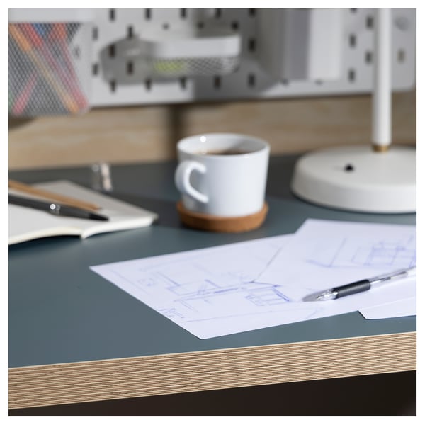 Desk with blue-green tabletop, white cup, sketch with pens, grey desk lamp, organiser in background.