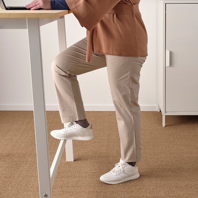 Person standing at tall table with footrest, wearing casual attire and white trainers.