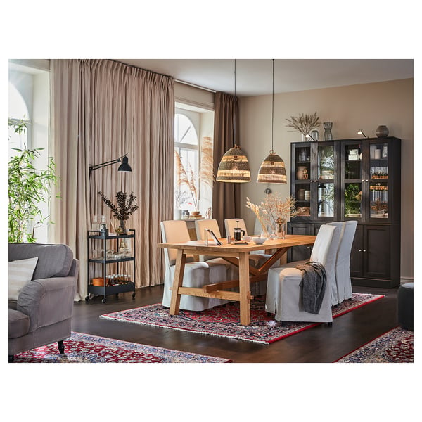 Rustic dining room with wooden table, grey chairs, black cabinet, and wooden pendants. Pink curtains, colourful rug, and plants add warmth.