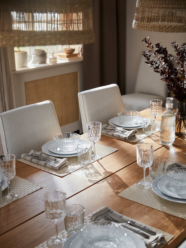 Modern dining table with white chairs, clear glassware, and plates. Neutral tones, natural wood, and elegant design.