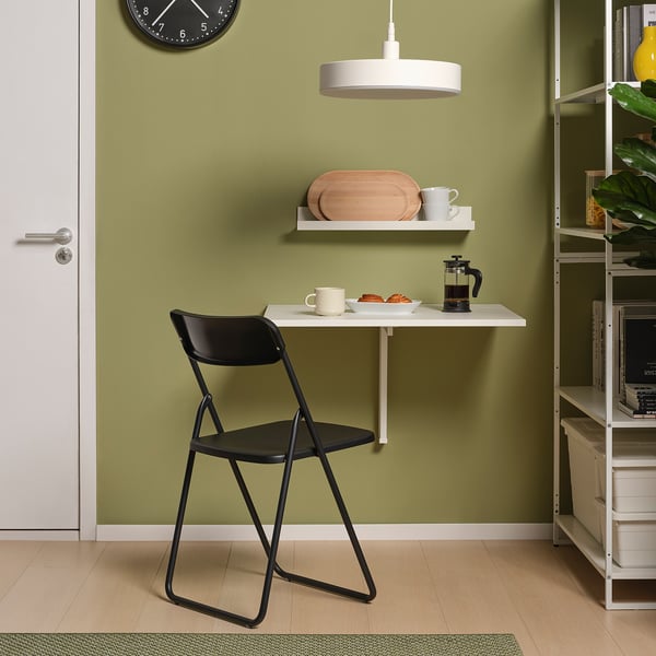 Small, wall-mounted white table, with black folding chair. Modern design, green wall, hanging clock and plant.