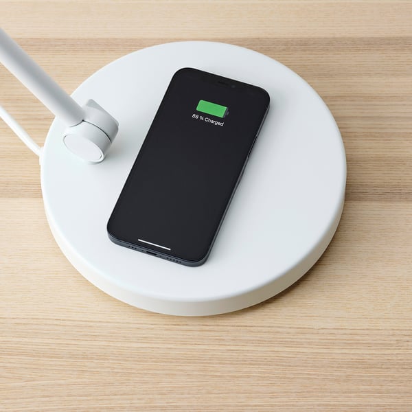 A smartphone wirelessly charging on a round, white NYMÅNE charging pad with a usb port and adjustable lamp.