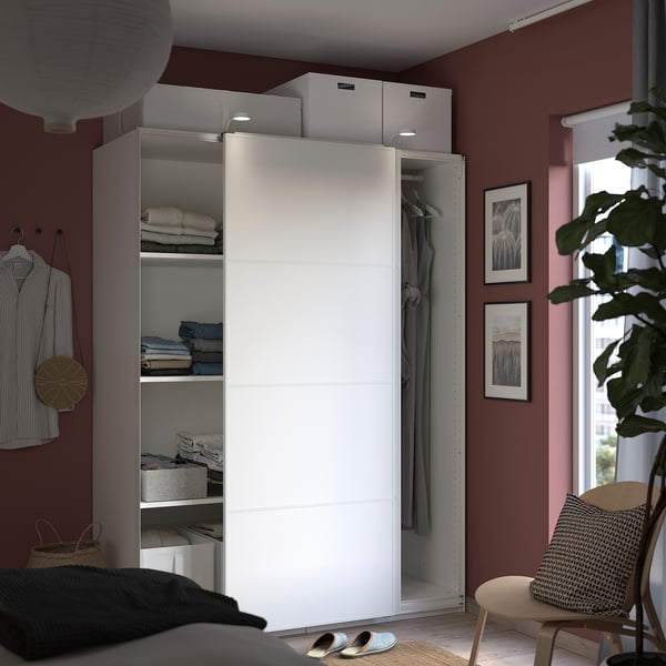 Modern white PAX wardrobe against red wall, open showing clothes and shelves, with a chair and plant beside it.