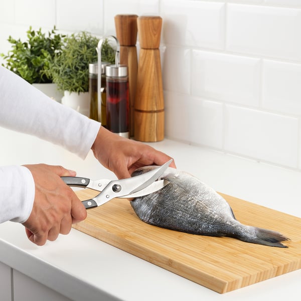 A person uses shears to cut fish on a wooden board in a kitchen. The shears have a locking mechanism for safe storage.