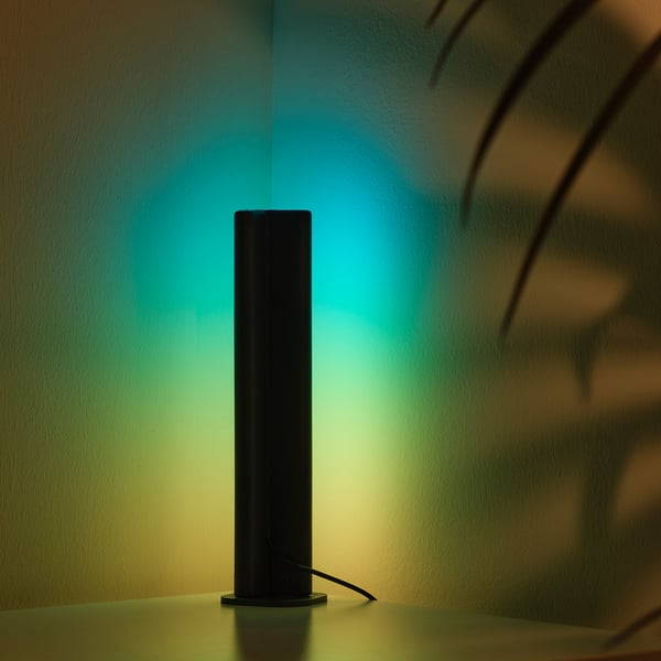 Black SKAFTSÄRV lamp on table, casting blue-green light.