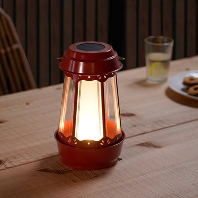 Red SOLVINDEN solar lamp on table beside drink & cookies; warm, glowing light, compact size.