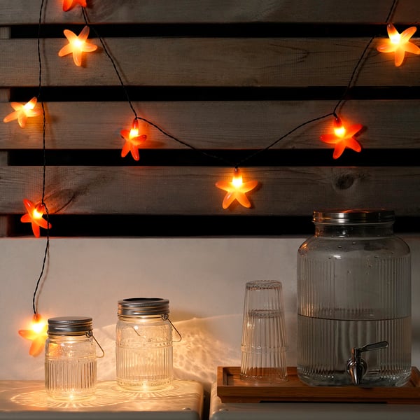 Multiple glass jars and drinks dispenser beneath warm orange star-shaped lights. Jars and dispenser appear small compared to large wooden wall slats.