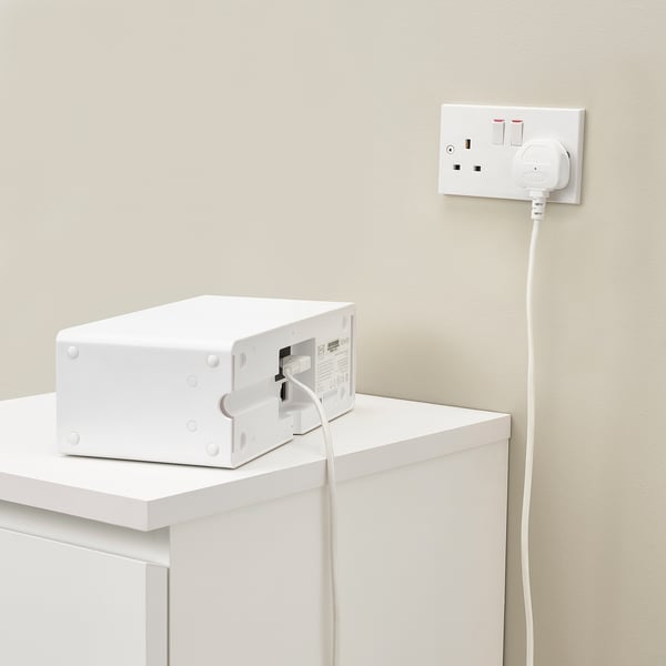 White speaker on white shelf, plugged into wall socket with long cord.