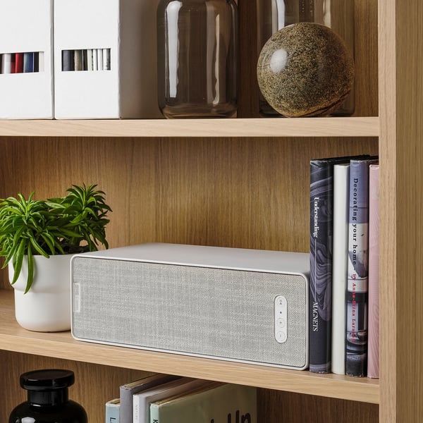 A white SYMFONISK wifi speaker on a wooden shelf playing music, paired with books and a plant.